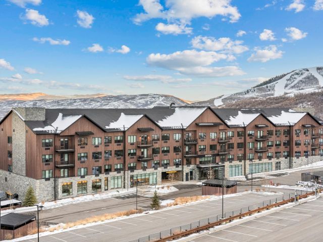 A large, multi-story brown building with balconies and a stone facade, set against a backdrop of snowy mountains and a blue sky with clouds.