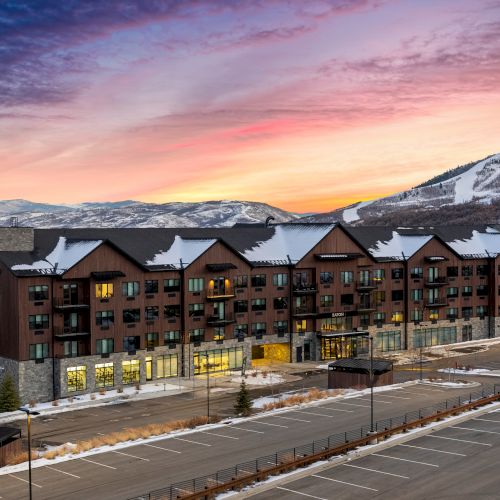 A large lodge-like building with snow-covered roofs, set against a colorful sunset, mountains, and an empty parking lot in the foreground.