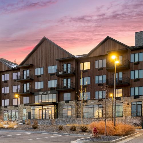 The image shows a modern, multi-story building with large windows and a stone facade, set against a colorful sunset sky.