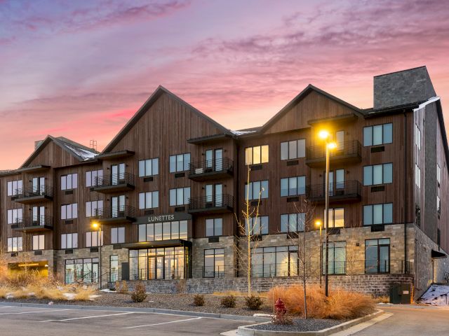 A multi-story building with a brown and stone exterior, a parking lot in front, and streetlights, set against a colorful sky.