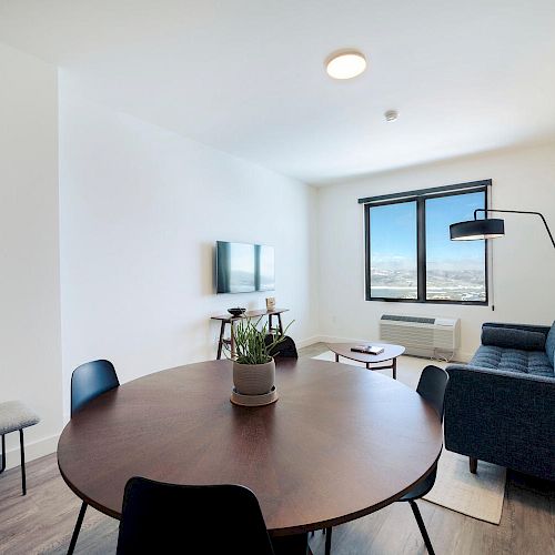 A modern living room with a round wooden table, black chairs, a gray sofa, a mounted TV, and a window with a city view.