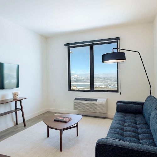 A modern living room with a blue sofa, coffee table, wall-mounted TV, and a large window with a scenic view.