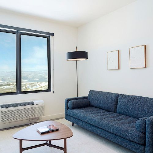 A modern living room features a blue sofa, a coffee table with books, a floor lamp, two framed pictures, and a large window with a scenic view.