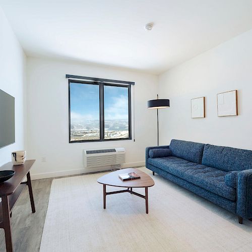 A modern, minimalist living room with a blue sofa, coffee table, wall art, TV, and a large window for natural light.