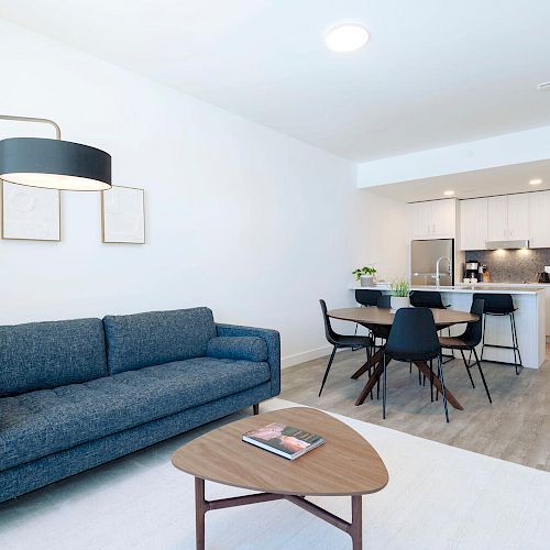 A modern living space with a blue couch, coffee table, and open kitchen area featuring a dining table and chairs. Minimalist decor.