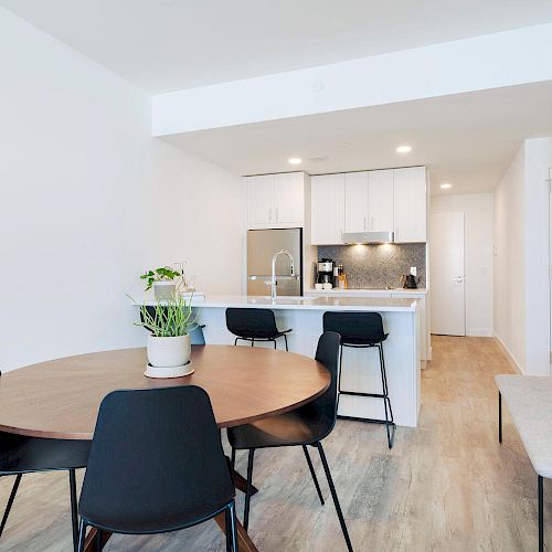 A modern kitchen and dining area with a round table, black chairs, a plant centerpiece, a counter with stools, and sleek white cabinetry.