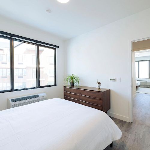 A clean, modern bedroom with a white bed, wooden dresser, large window, and a doorway leading to another room with similar decor and flooring.