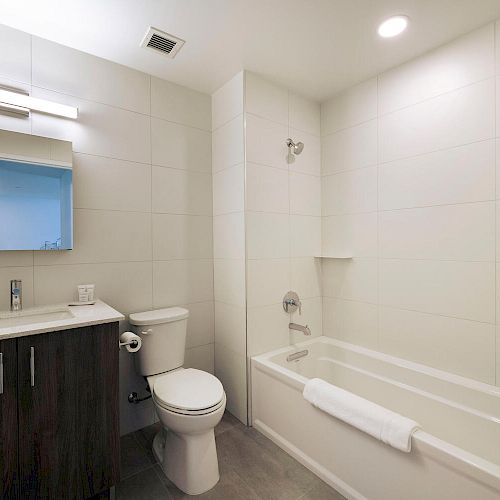 This image shows a modern bathroom with a bathtub, toilet, sink with a mirror above, and a cabinet below. The space is well-lit and clean.