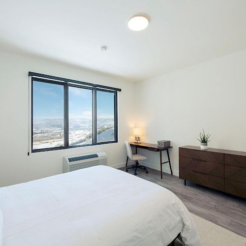 A minimalist bedroom with a large bed, a wooden dresser, a desk, a window showing a scenic view, and simple decor.