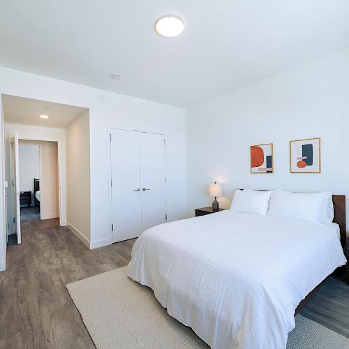 A modern bedroom with a white bed, two nightstands with lamps, a dresser, and abstract artwork on the wall. White walls and wooden floors.