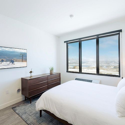 A bedroom with a bed, dresser, lamp, and wall art, featuring large windows with a scenic view. The room is bright and minimalist in design.