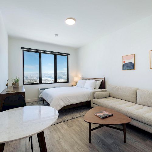 A modern bedroom features a bed, a couch, a small coffee table, and a round dining table. Two abstract artworks hang on the wall.