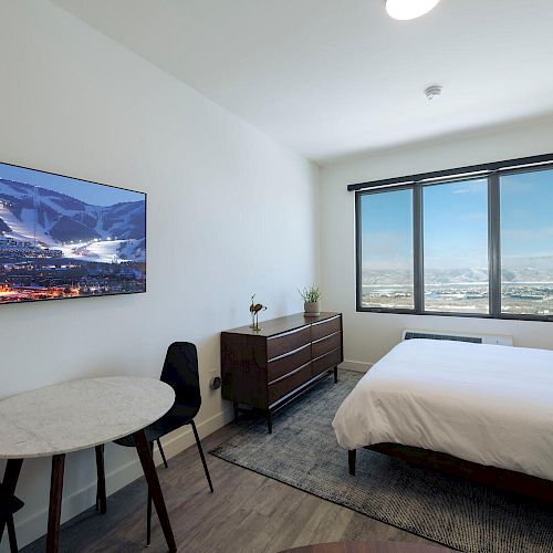 A modern hotel room with a large window, a bed, a TV displaying a snowy landscape, a small table with chairs, and a dresser.