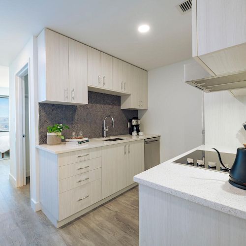 A modern kitchen with white cabinetry, a sink, stove, and kettle. The kitchen leads to a sitting area with a table and chairs near a window.