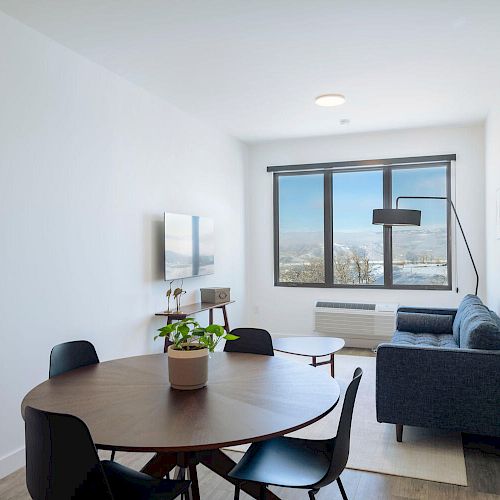 A modern living room with a round dining table, four chairs, a grey couch, a wall-mounted TV, and a large window with a view.