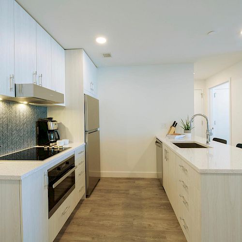 A modern kitchen with a stove, oven, refrigerator, sink, and an island counter with cabinets. The backsplash has a tile design and the floor is wooden.