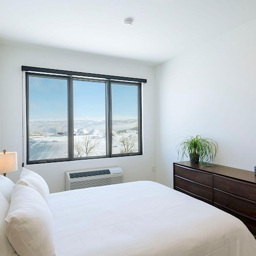 A minimalist bedroom with a large window, white bed, wooden dresser, potted plant, and a lamp, all overlooking a scenic view.