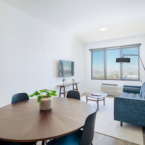 Modern living room with a round dining table, potted plant, blue sofa, wall-mounted TV, floor lamp, and large window with city view.