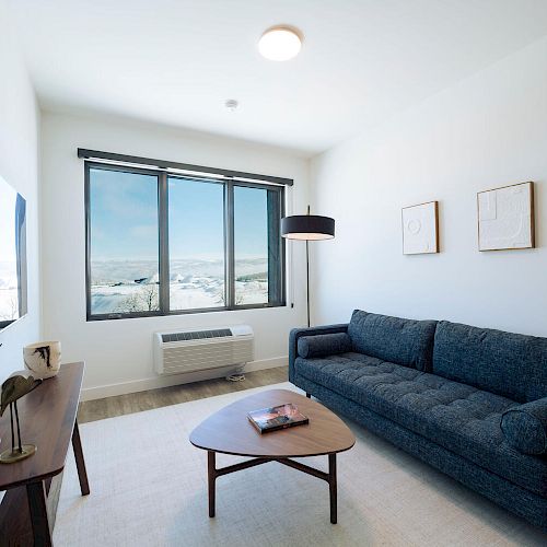 A minimalist living room with a blue couch, coffee table, TV, lamp, and large window with a scenic view, plus simple wall art.