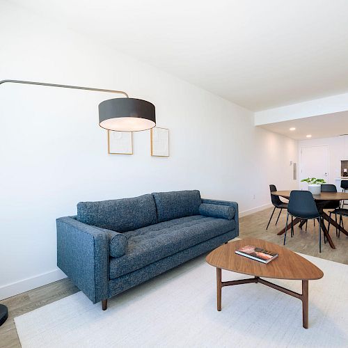 A modern living room with a gray sofa, floor lamp, wooden coffee table, open dining area with black chairs, and adjacent white kitchen.