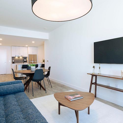 The image shows a modern living room with a blue sofa, a wooden coffee table, a large TV on the wall, and a dining area in the background.