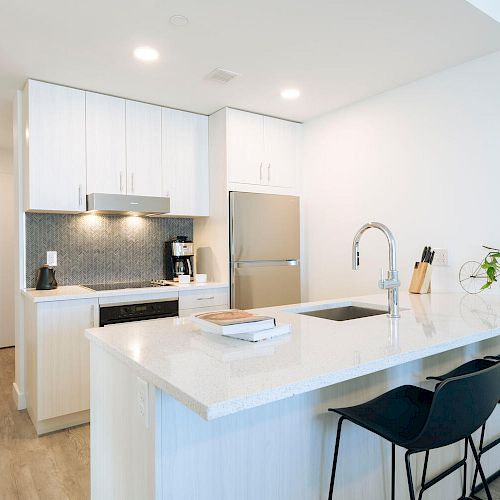 Modern kitchen with white cabinets, stainless steel appliances, island with sink, three stools, and a small plant on the counter.