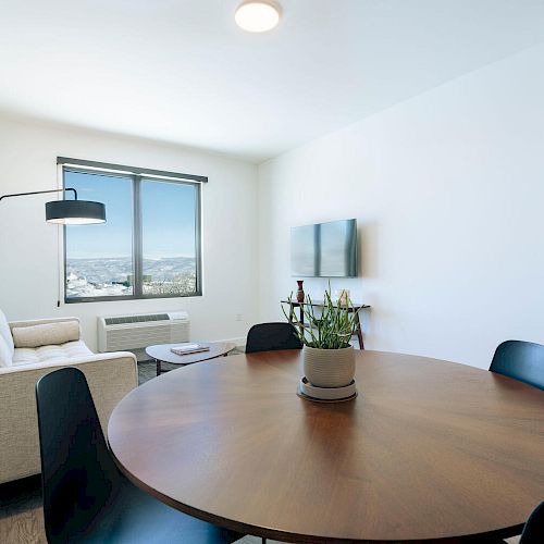 The image shows a modern, minimalistic living and dining area with a round table, black chairs, a beige sofa, wall-mounted TV, and a large window.