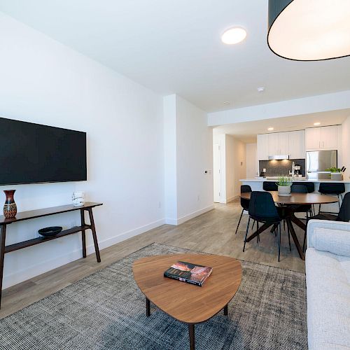 A modern living room with a wall-mounted TV, grey sofa, coffee table, dining area, and a kitchen in the background.