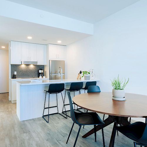 The image shows a modern kitchen and dining area with white cabinets, a round wooden table, black chairs, and a countertop bar with stools.