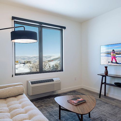 A cozy room with a white couch, a modern floor lamp, a window view, a TV mounted on the wall, and a wooden coffee table with a book.