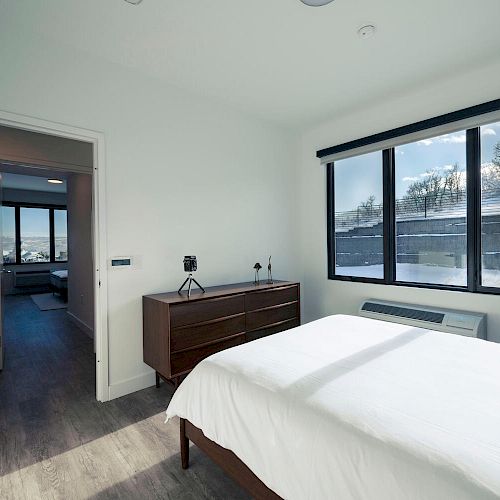 A bright bedroom with large windows, a bed with white linens, a wooden dresser, and an open door leading to another room. Snowy outdoor scenery.