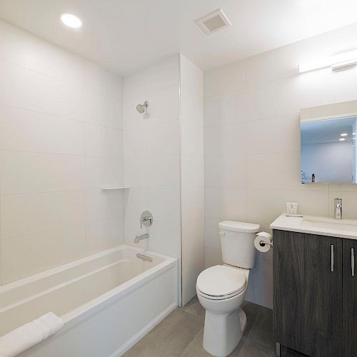 A modern bathroom with white walls, bathtub, toilet, and a vanity with a mirror, soap dispenser, and toothbrush holder on top.