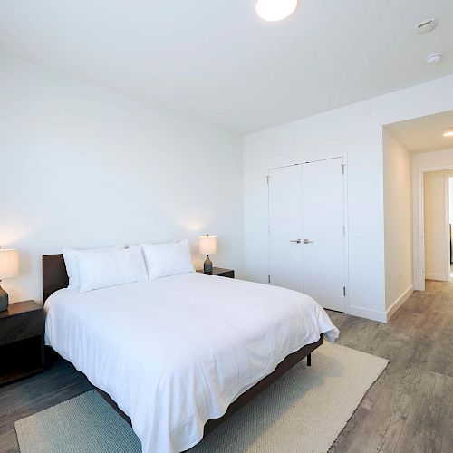 A minimalist bedroom features a neatly made bed with white linens, two bedside tables with lamps, and light wood flooring leading to a hallway.