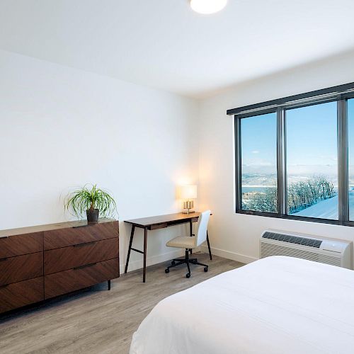 A minimalist bedroom featuring a bed, dresser with a plant, desk with a chair and lamp, and a large window with a scenic view.