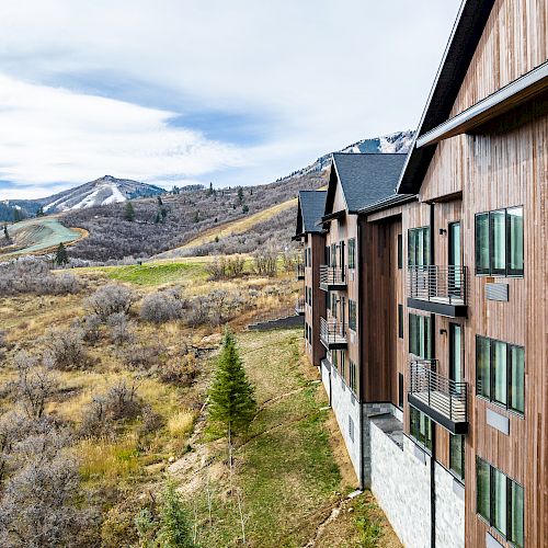 A large wooden building is perched on a hillside with mountainous terrain and sparse vegetation in the background under a partly cloudy sky.