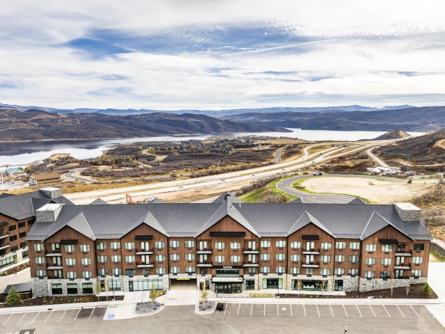 Aerial view of a multi-story brick building with a gray roof, a parking lot in front, and a scenic background of mountains and a lake.