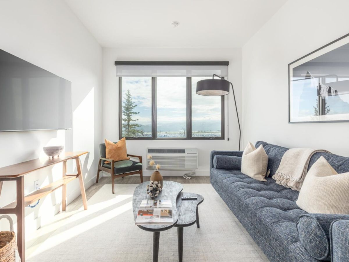 A modern living room with a grey couch, coffee table, large window, wall-mounted TV, and a bookshelf. A relaxing and stylish space.