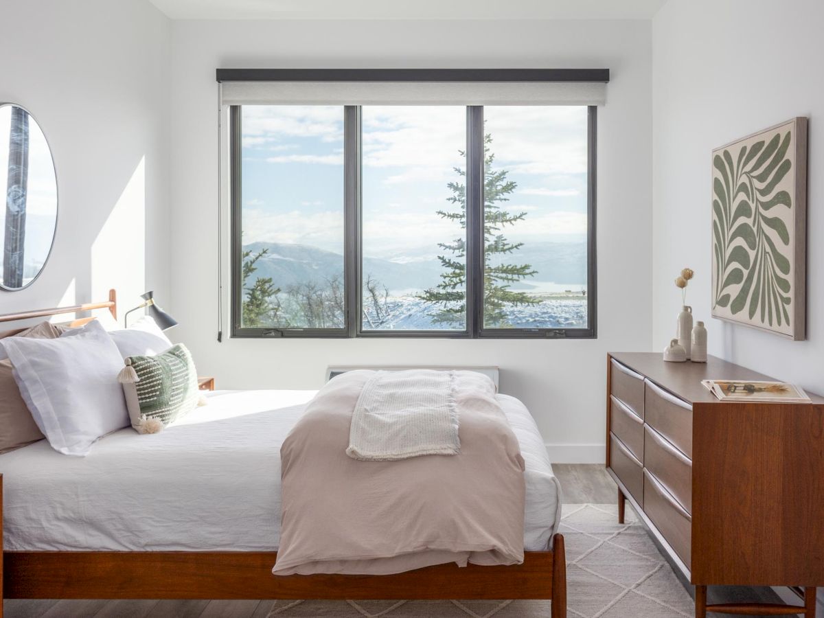 A modern bedroom with a large window overlooking a scenic view, featuring a bed, dresser, artwork, and a mirror on the wall.