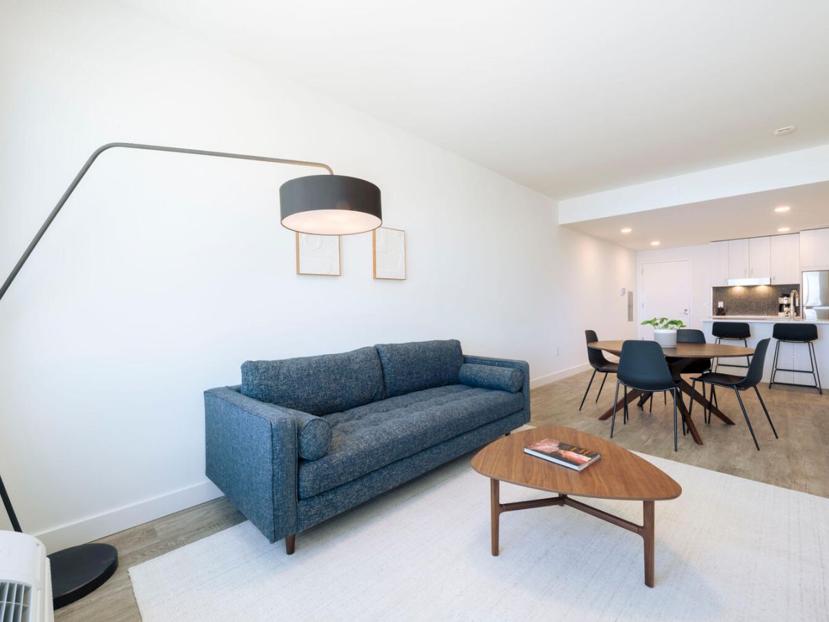 This image depicts a modern living room with a blue sofa, wooden coffee table, floor lamp, and a dining area with a kitchen in the background.