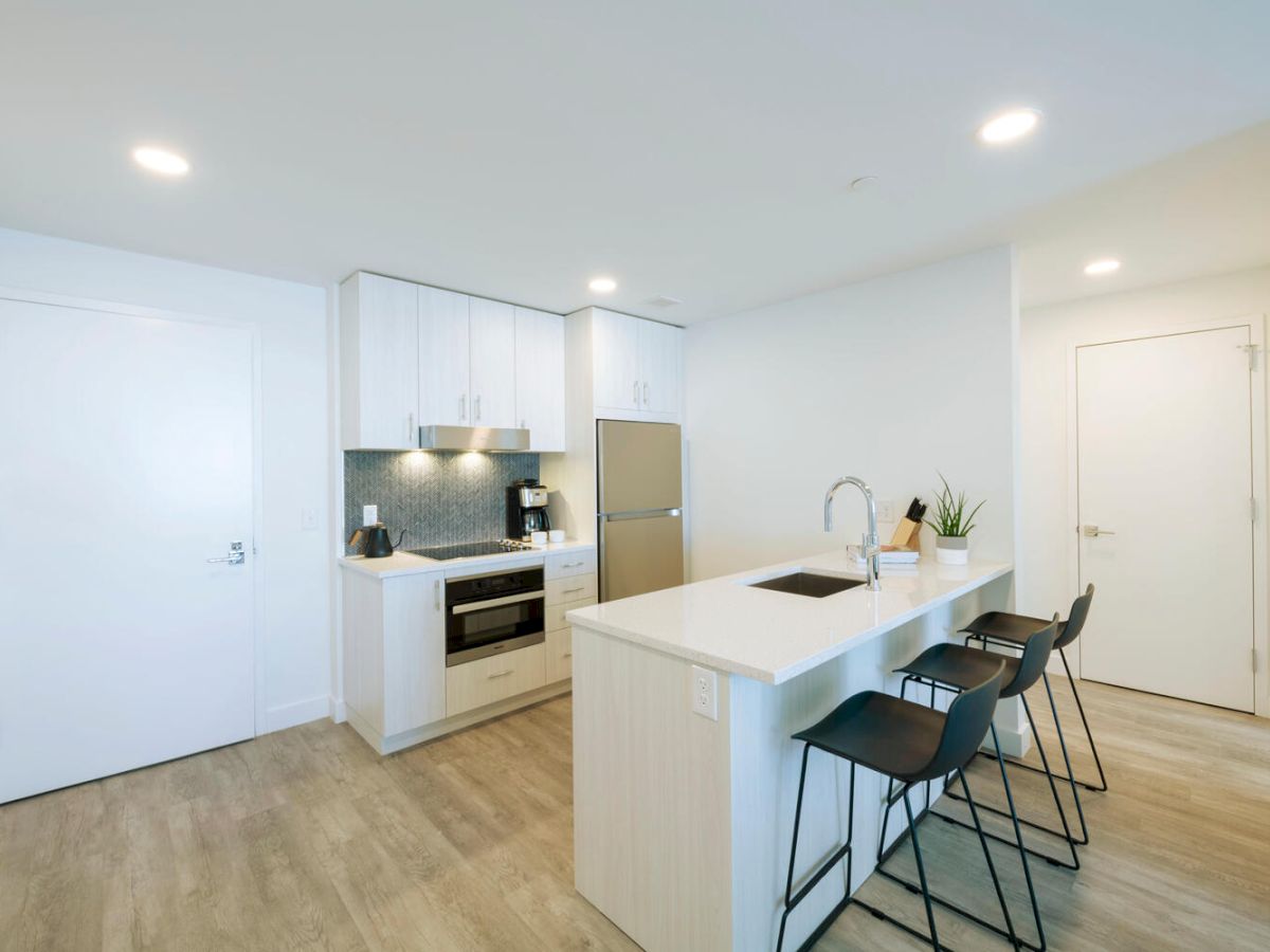 Modern kitchen with white cabinets, stainless steel appliances, island with sink, three black barstools, and wood flooring in compact space.