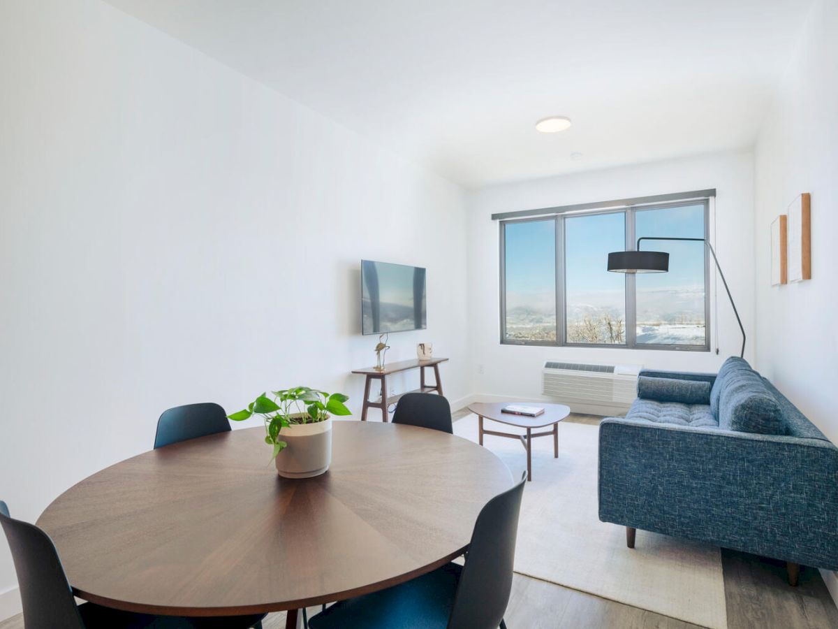 A modern living room with a round dining table, black chairs, blue couch, wall-mounted TV, standing lamp, and potted plant, featuring a large window.