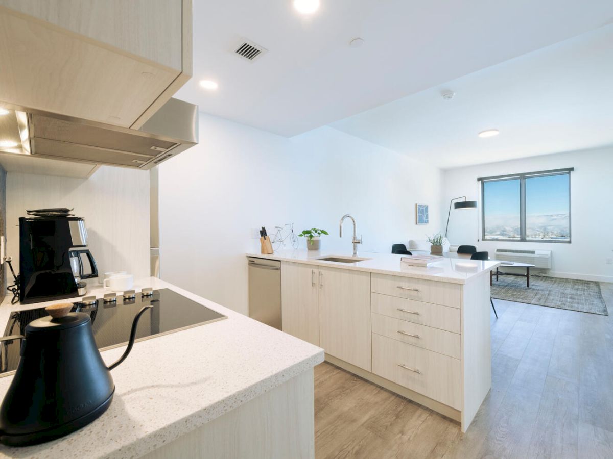 Modern kitchen with white cabinets, island, and appliances, leads to an open living area with large window and seating arrangement.