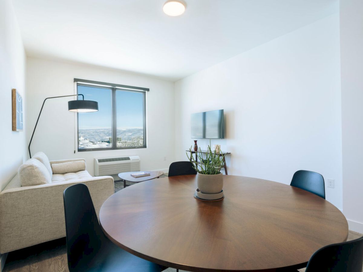 A modern, bright room with a round wooden table, four black chairs, a mounted TV, a white sofa, a tall lamp, a window, and a small plant.