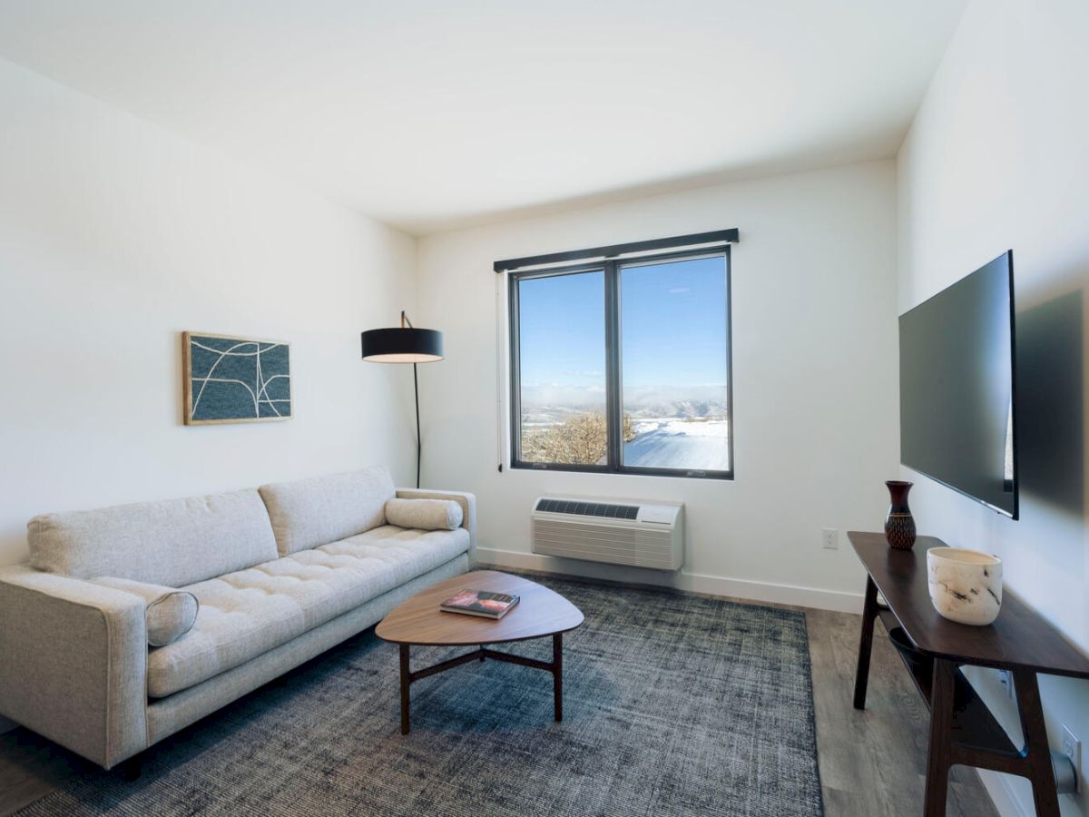 A modern living room with a white sofa, coffee table, wall-mounted TV, window with blinds, floor lamp, wall art, and a side table with decor items.