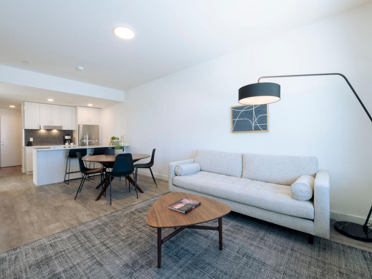 A modern living room features a gray couch, wooden coffee table, and a black floor lamp, all adjacent to a kitchen with a dining area.