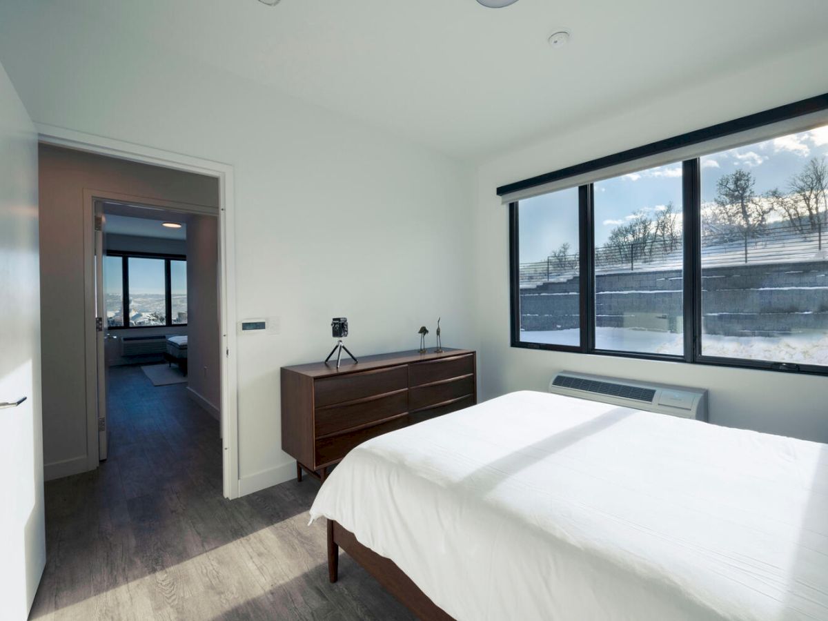 A minimalist bedroom with a large window showcasing a snowy landscape, a wooden dresser, white bed, and a door leading to another room.