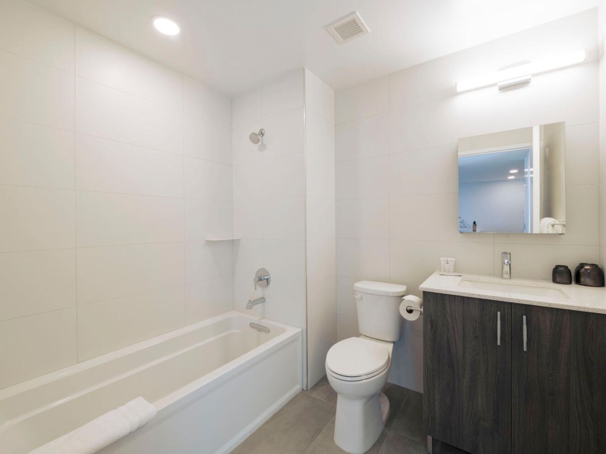 The image shows a modern bathroom with a bathtub, toilet, vanity with a sink, and a mirror above the sink. The design is minimalist.