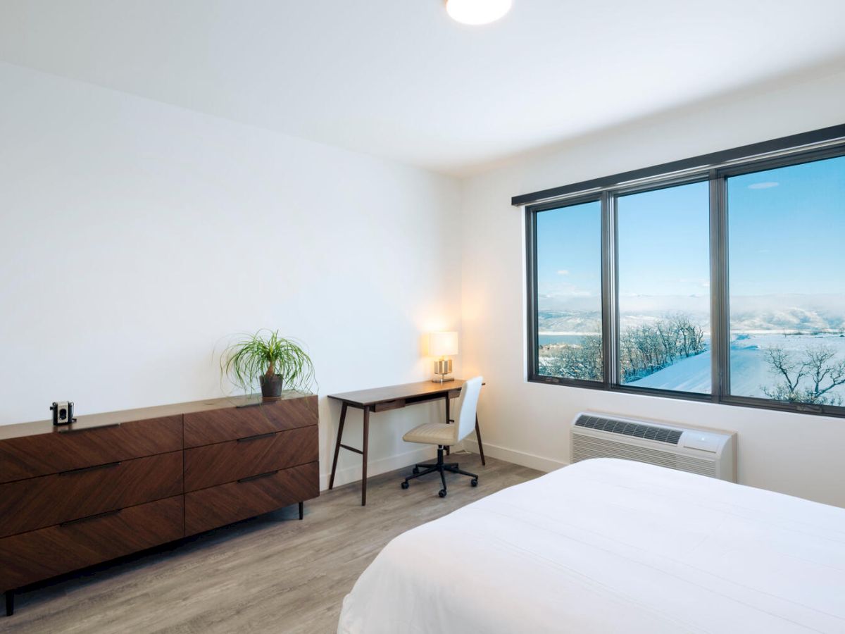 A minimalistic bedroom with a bed, dresser, desk, and chair. The room has a large window with a scenic view and a potted plant on the dresser.