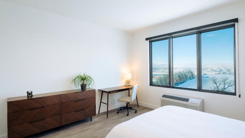 A hotel room with a bed, desk, dresser, and a window showing a snowy landscape.