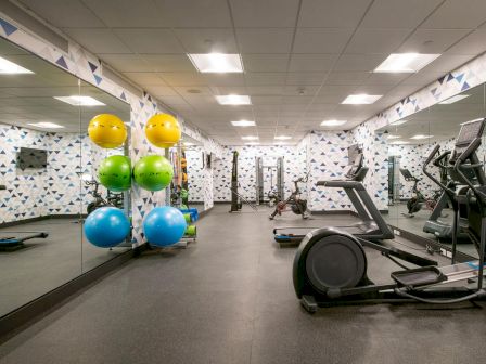 A gym with various equipment including an elliptical machine, exercise ball rack with colorful balls, mirrored wall, and patterned wallpaper.